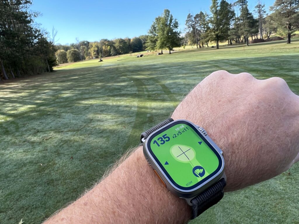 Golfer's wrist with an Apple Watch Ultra and a golf hole in the background. The sky is blue and the golfer appears to be in the middle of the fairway. The watch shows a map of the hole and indicates that the center of the green is 135 yards away. It also indicates that it's hole #2, a par 5, handicap 11.